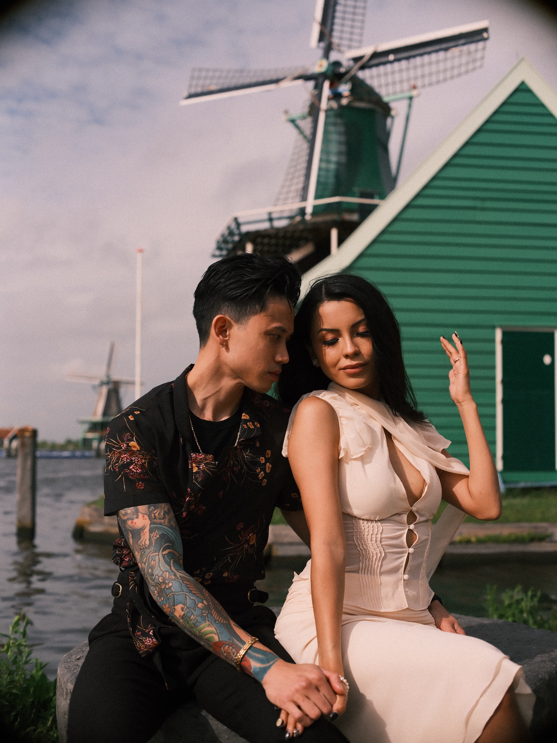 Woman with windblown dark hair raising one hand as she sits beside her partner on a millstone, a green house and windmill behind