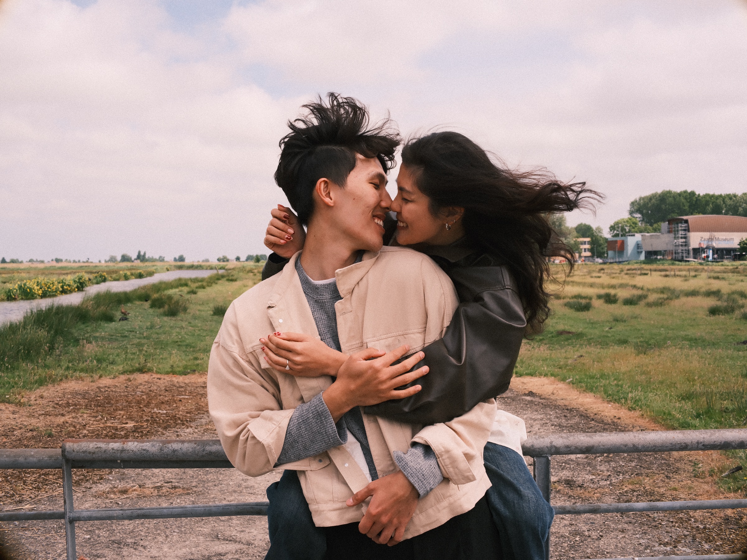 Couple kissing at a metal farm gate while the wind blows her long hair sideways, flat green fields behind the Zaanse Schans