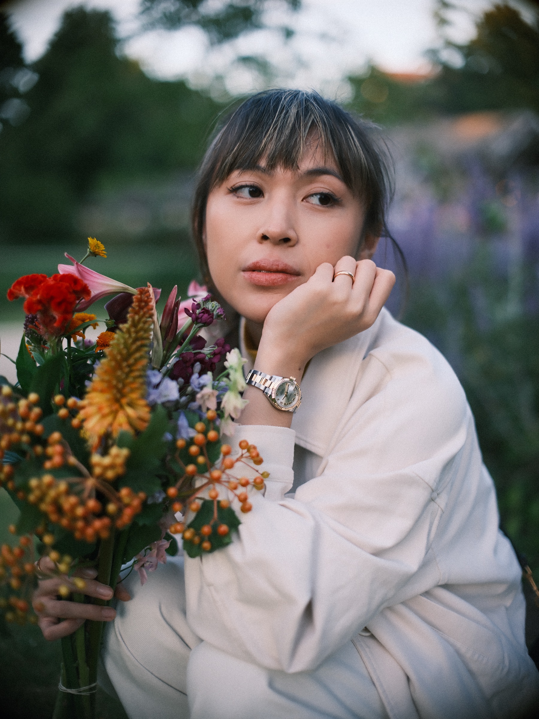 Vrouw in een crèmekleurige jas met haar kin op haar hand naast een bos rode en oranje bloemen, paarse bloemen zacht erachter
