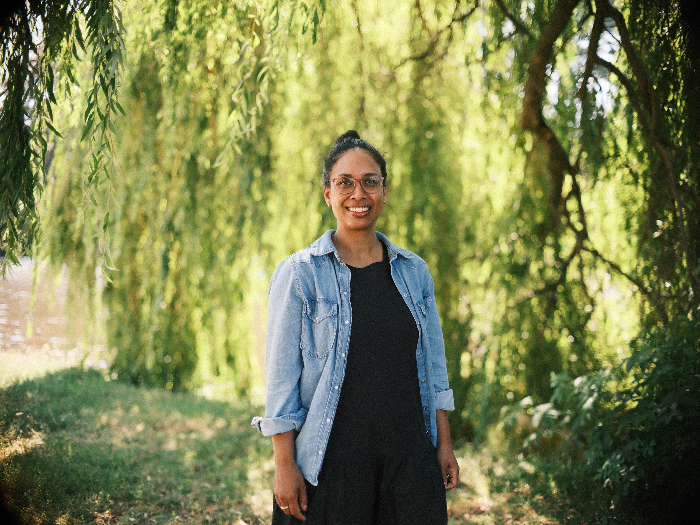 Vrouw met bril en spijkerblouse lacht onder een treurwilg in Leiden, met water tussen de takken op een zomerse middag