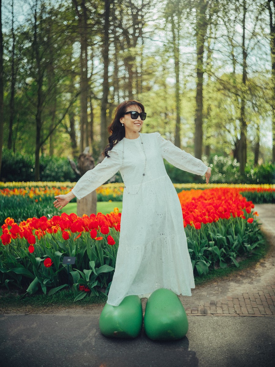Vrouw met zonnebril en lange witte jurk poseert met wijde armen voor reusachtige groene klompen in Keukenhof