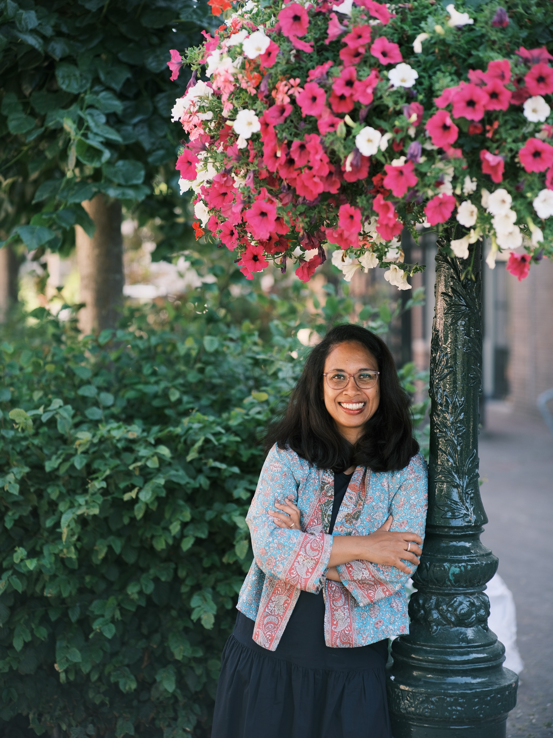 Lachende vrouw in een jasje met paisleyprint leunt tegen een donkergroene lantaarnpaal onder een hangmand met roze en witte petunia’s in Leiden