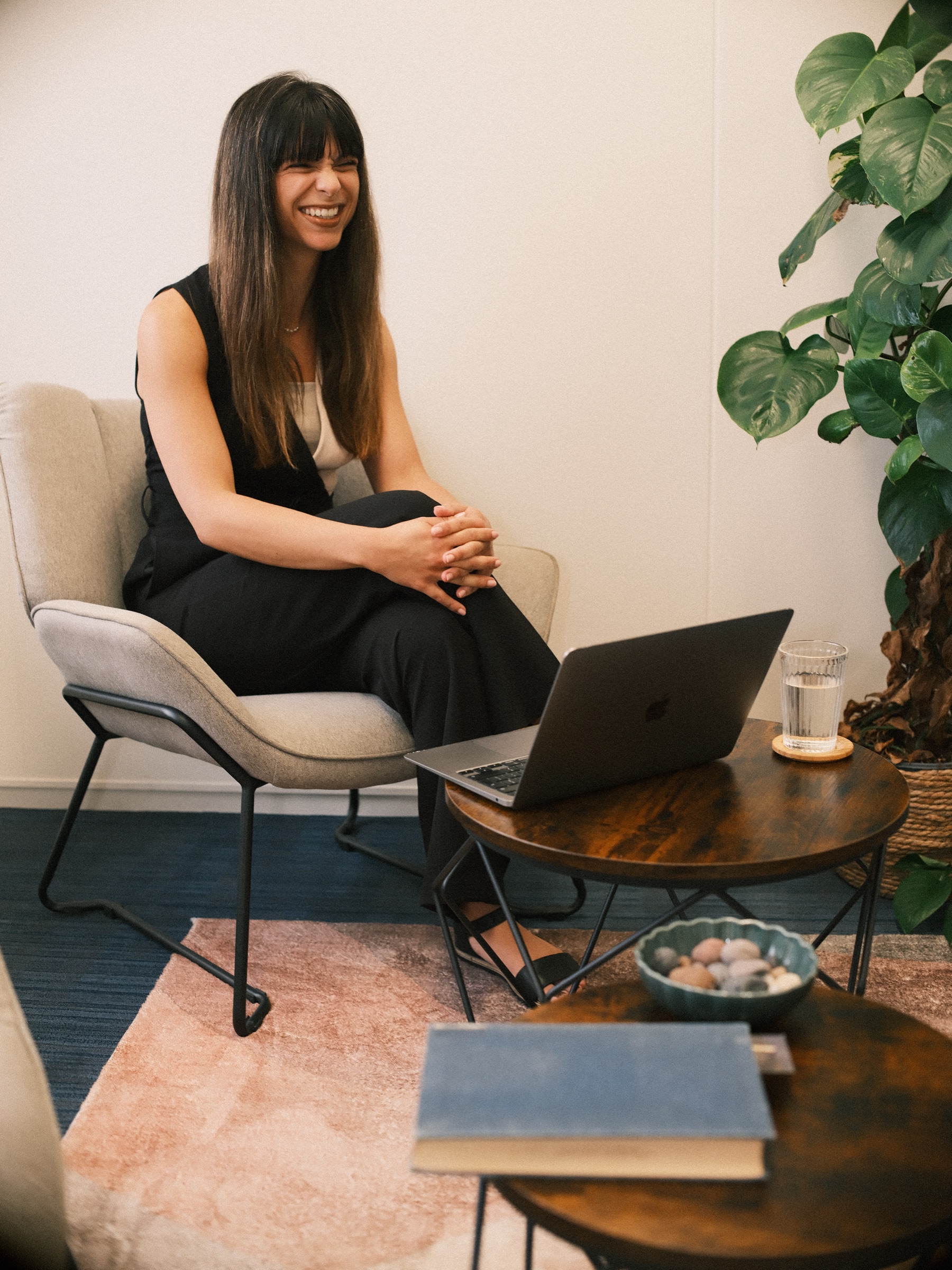 Woman in black laughing in a gray armchair with her laptop open, a large monstera and a glass of water beside her