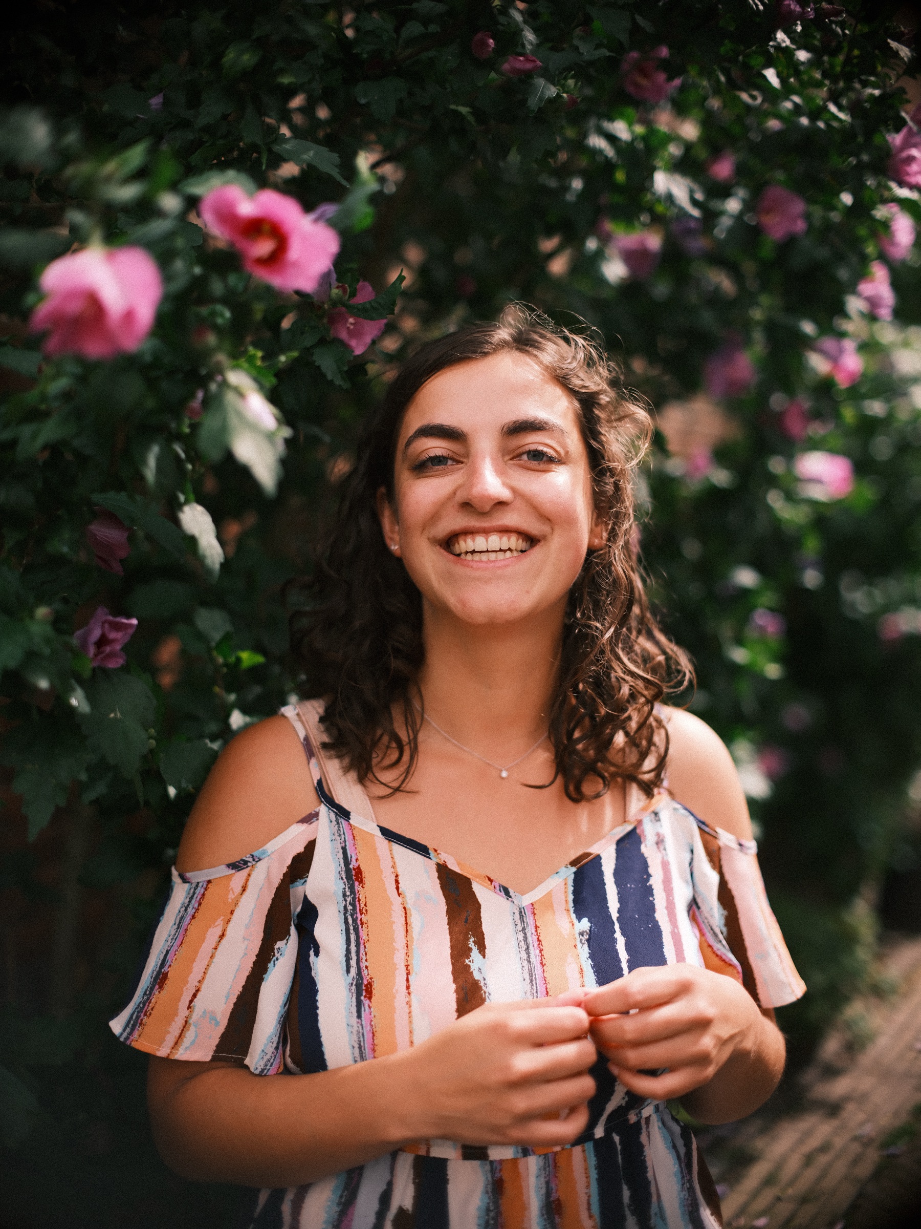 Vrouw lacht voor een struik vol roze bloemen in de zomer, de handen samen voor haar gestreepte jumpsuit