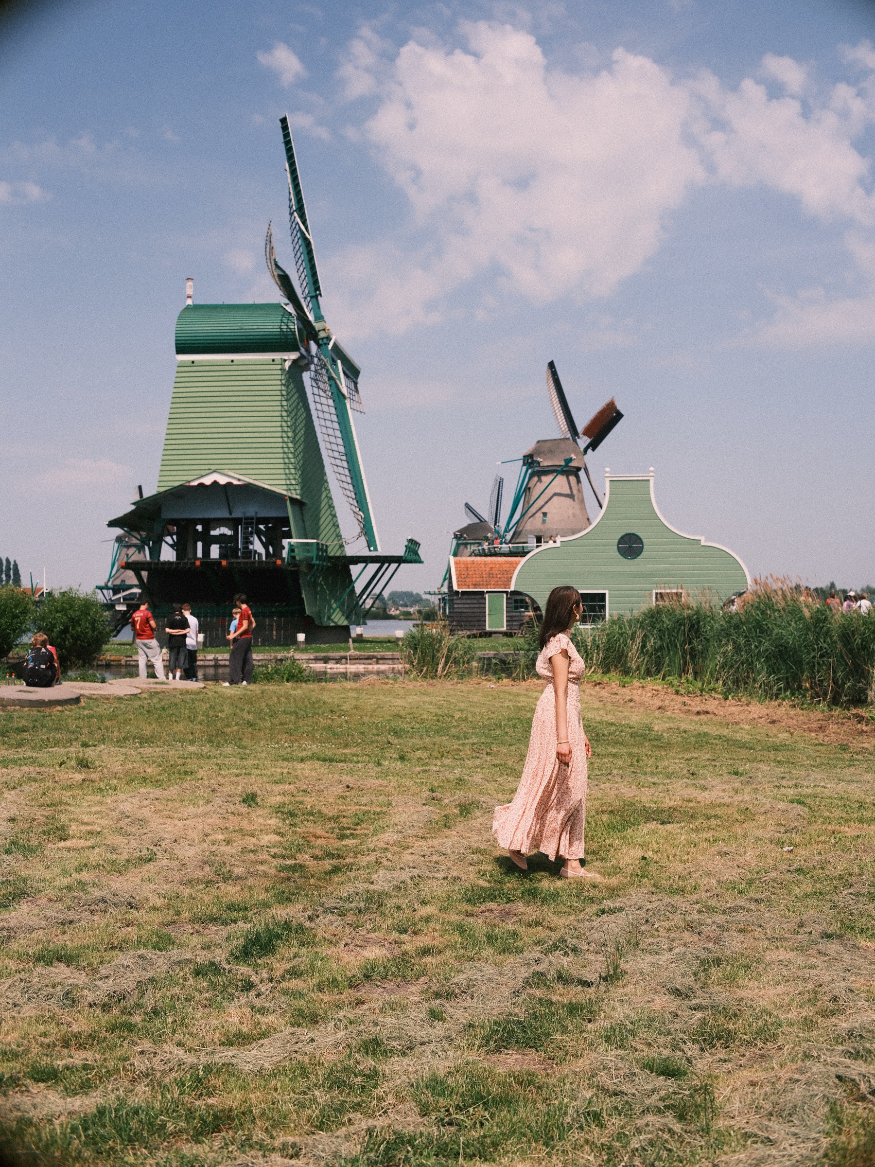 Woman in a long pink dress walking across a sunny meadow, visitors gathered around a big green mill and a light green gabled house behind
