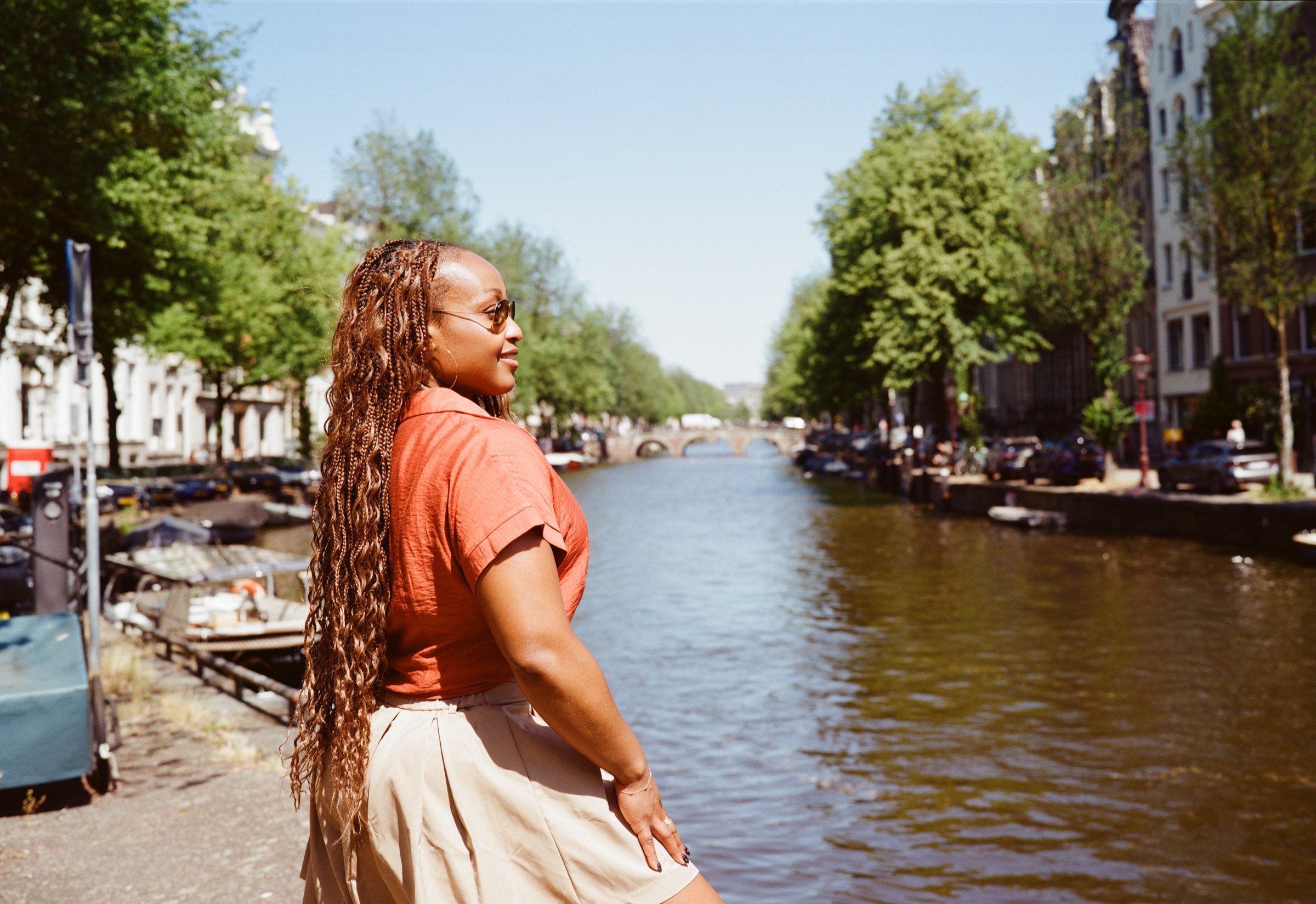 Profile of a woman in an orange shirt and beige shorts by a wide tree-lined canal in Amsterdam on a sunny summer day