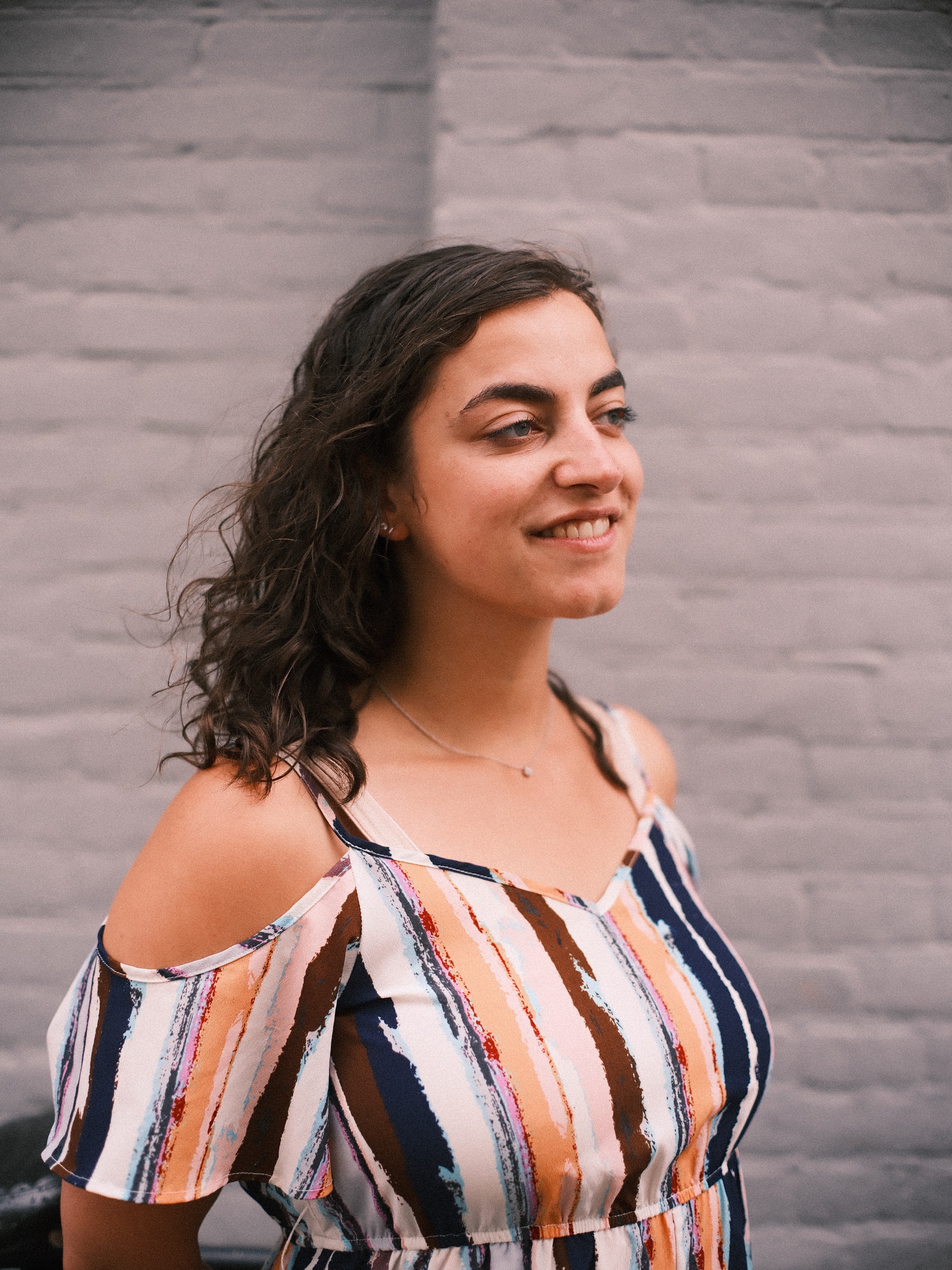Vrouw met donkere krullen glimlacht opzij voor een witgeverfde bakstenen muur, haar gestreepte top laat de schouders vrij