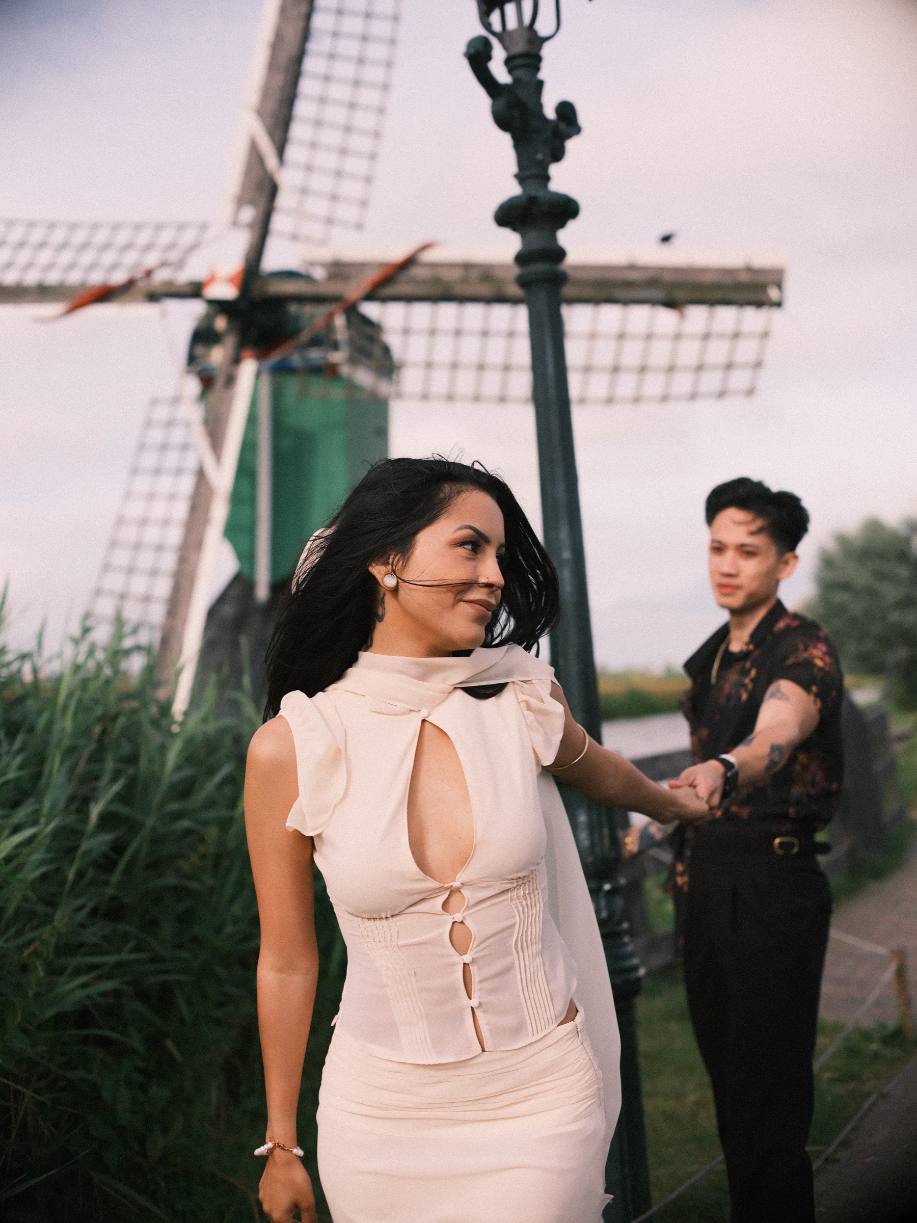 Woman in an ivory two-piece spinning away from her partner while he holds her hand, beside tall reeds and a small windmill