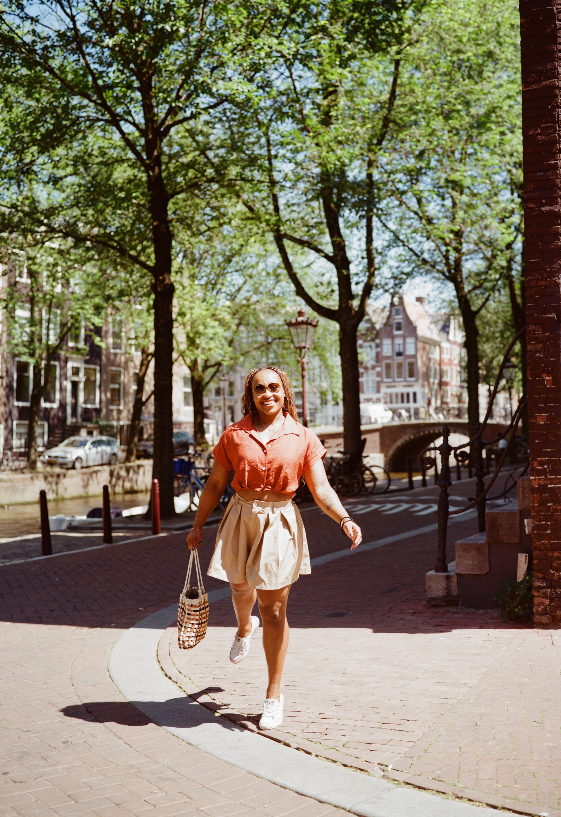 Vrouw in een beige plooishort en witte sneakers steekt met een gevlochten tas een klinkerstraat over, met bomen en een brug erachter