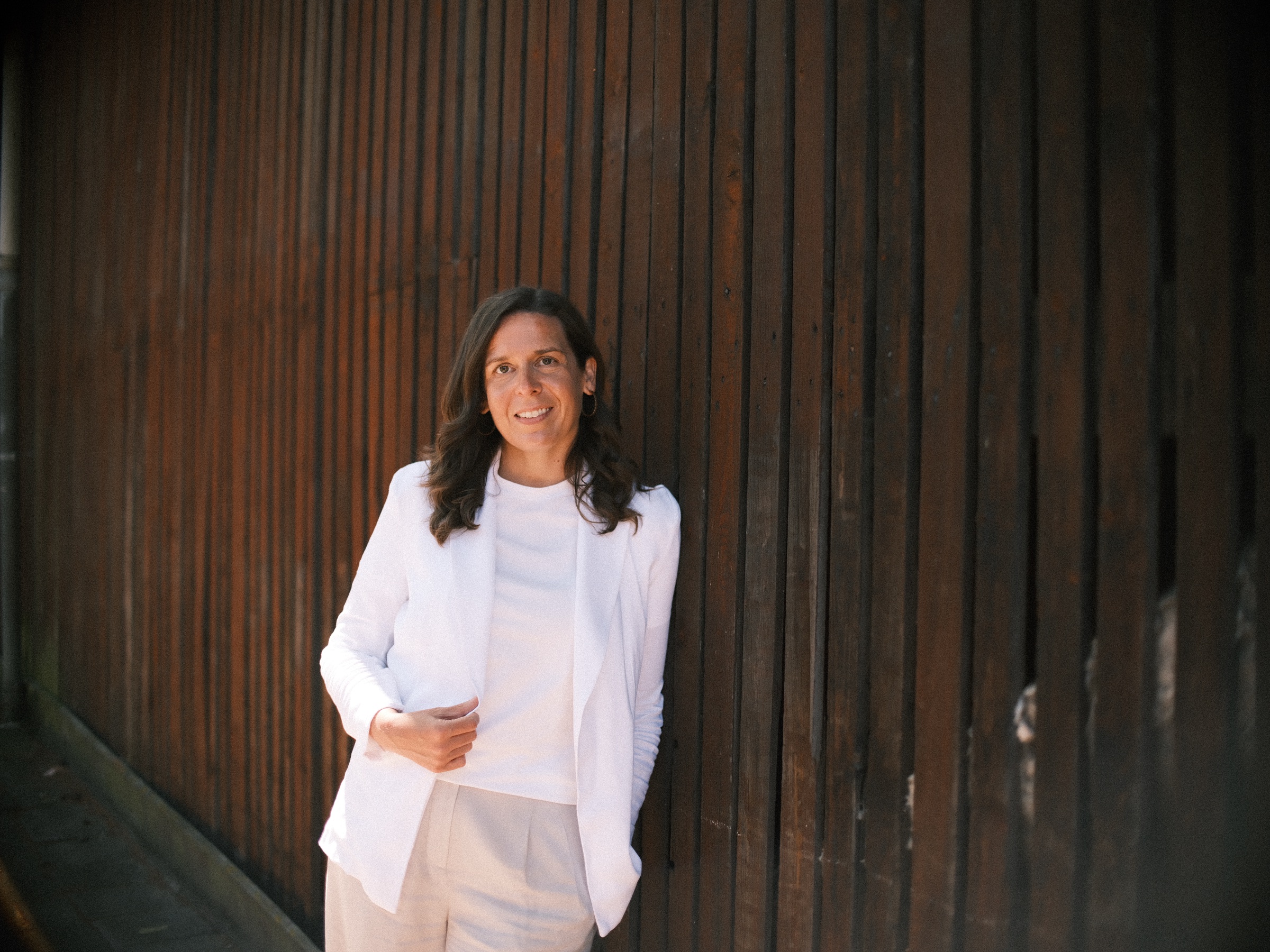 Vrouw in een witte blazer leunt lachend tegen een wand van donkere verticale planken, tijdens een solo fotoshoot in Haarlem