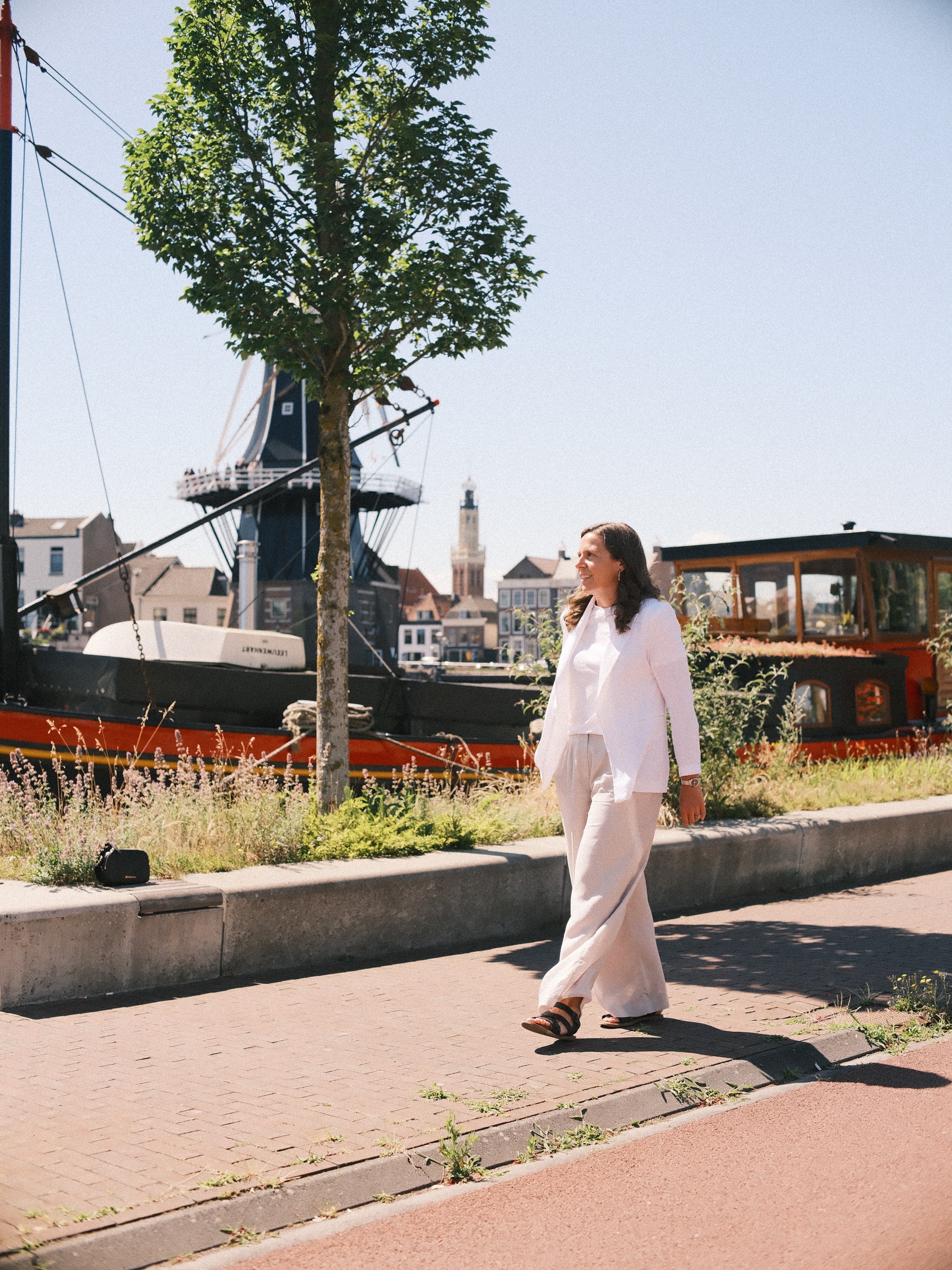 Vrouw in een witte blazer en wijde lichte broek loopt langs het Spaarne tijdens een solo fotoshoot, met molen De Adriaan en een oude schuit achter haar