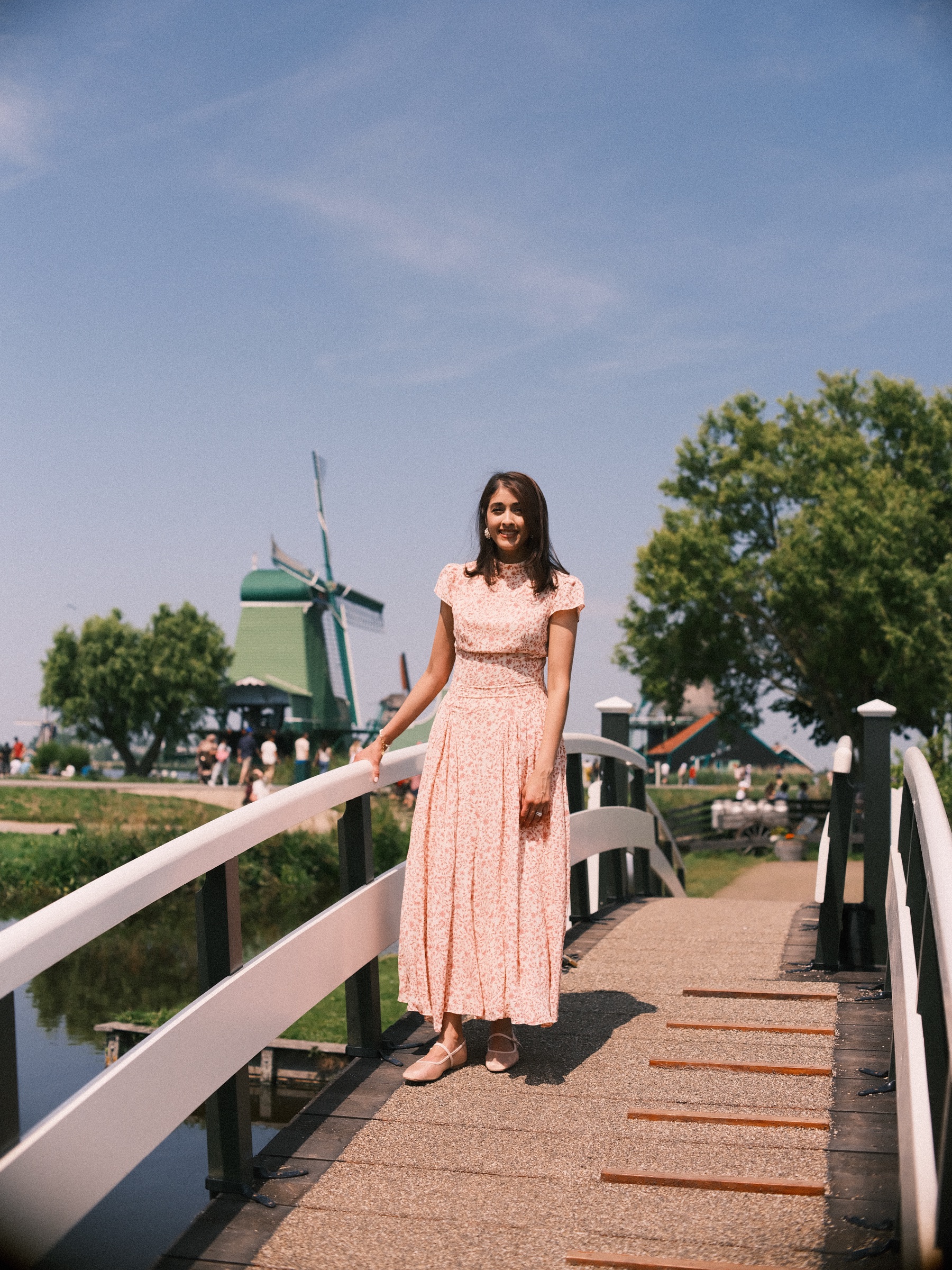 Woman in a long pink floral dress standing on a small white wooden bridge at the Zaanse Schans, a green mill and trees behind her in the sun