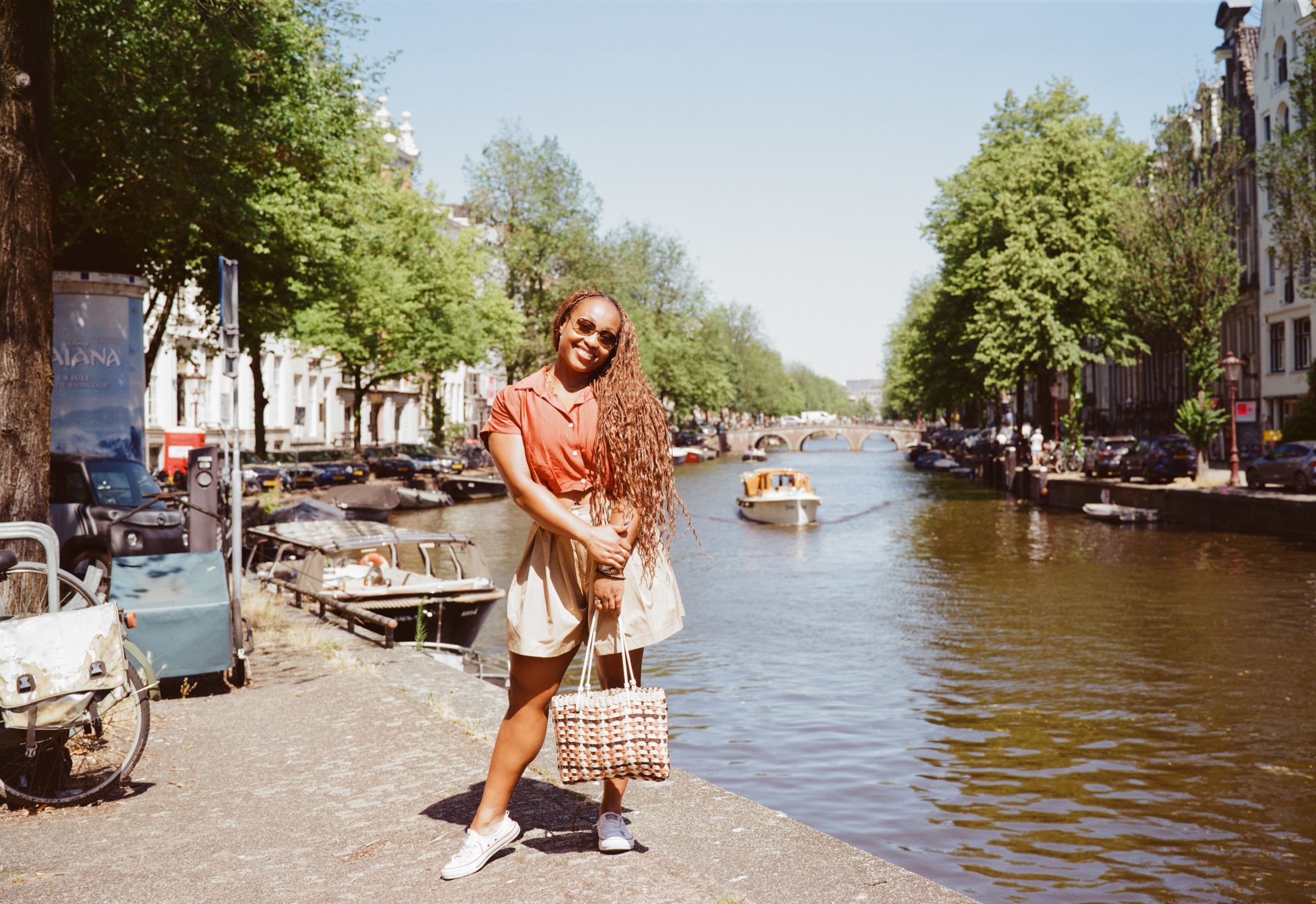 Smiling woman holding a woven bag on a canal quay in Amsterdam while a small boat comes up the water behind her